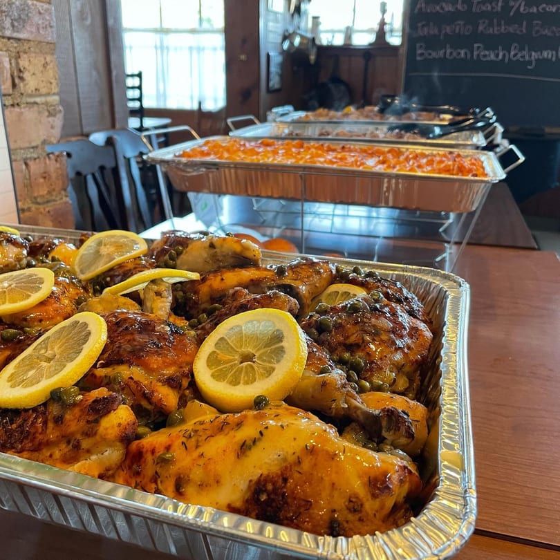 Lemon-herb roasted chicken in a foil pan, with other buffet dishes and a chalkboard menu in the background.
