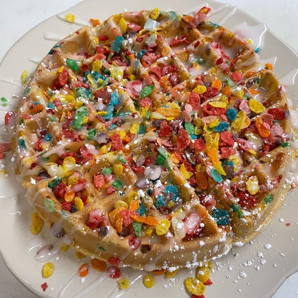 A waffle topped with colorful fruity cereal, white icing, and powdered sugar on a white plate.