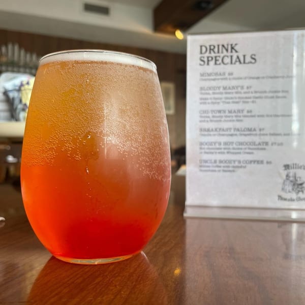 A vibrant orange and red drink in a stemless glass sits on a wooden table next to a drink specials menu.