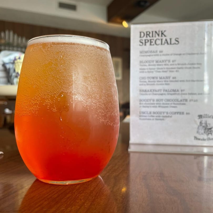 A vibrant orange and red drink in a stemless glass sits on a wooden table next to a drink specials menu.