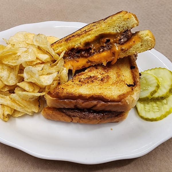 Cheeseburger melt on toasted bread with melted cheese, served with potato chips and pickle slices.
