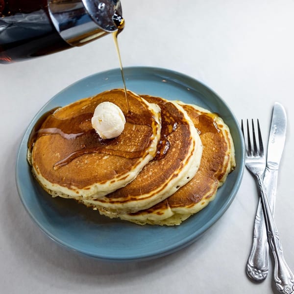 Maple syrup being poured over a stack of pancakes with butter on a blue plate.