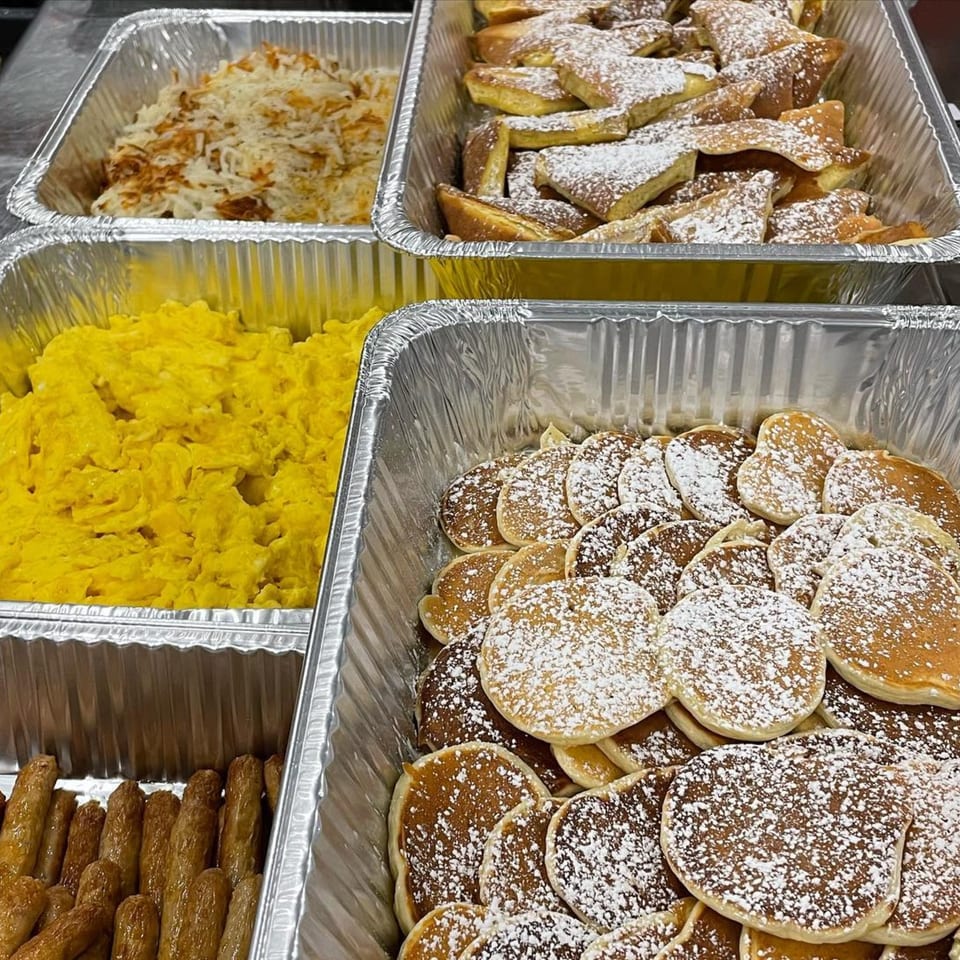 Catering trays filled with scrambled eggs, hash browns, French toast, mini pancakes, and sausage links.