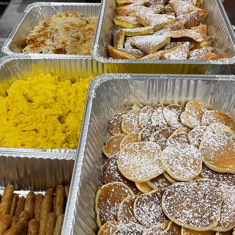 Catering trays filled with scrambled eggs, hash browns, French toast, mini pancakes, and sausage links.