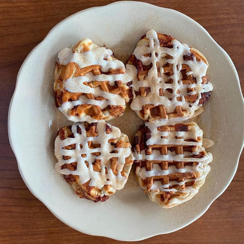 Four cinnamon roll waffles drizzled with white icing on a light-colored plate.